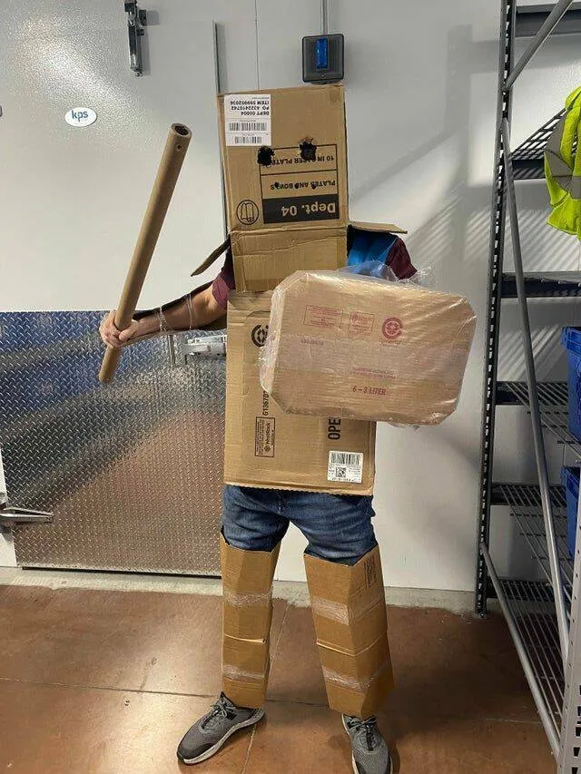 Osoba oblečená v kartónovom brnení s kartónovým štítom a papierovou rolkou ako mečom stojí v sklade.