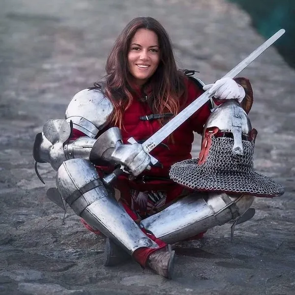 Žena v rytierskom brnení sedí s mečom, prilbou a úsmevom na tvári, stredoveká tematika.