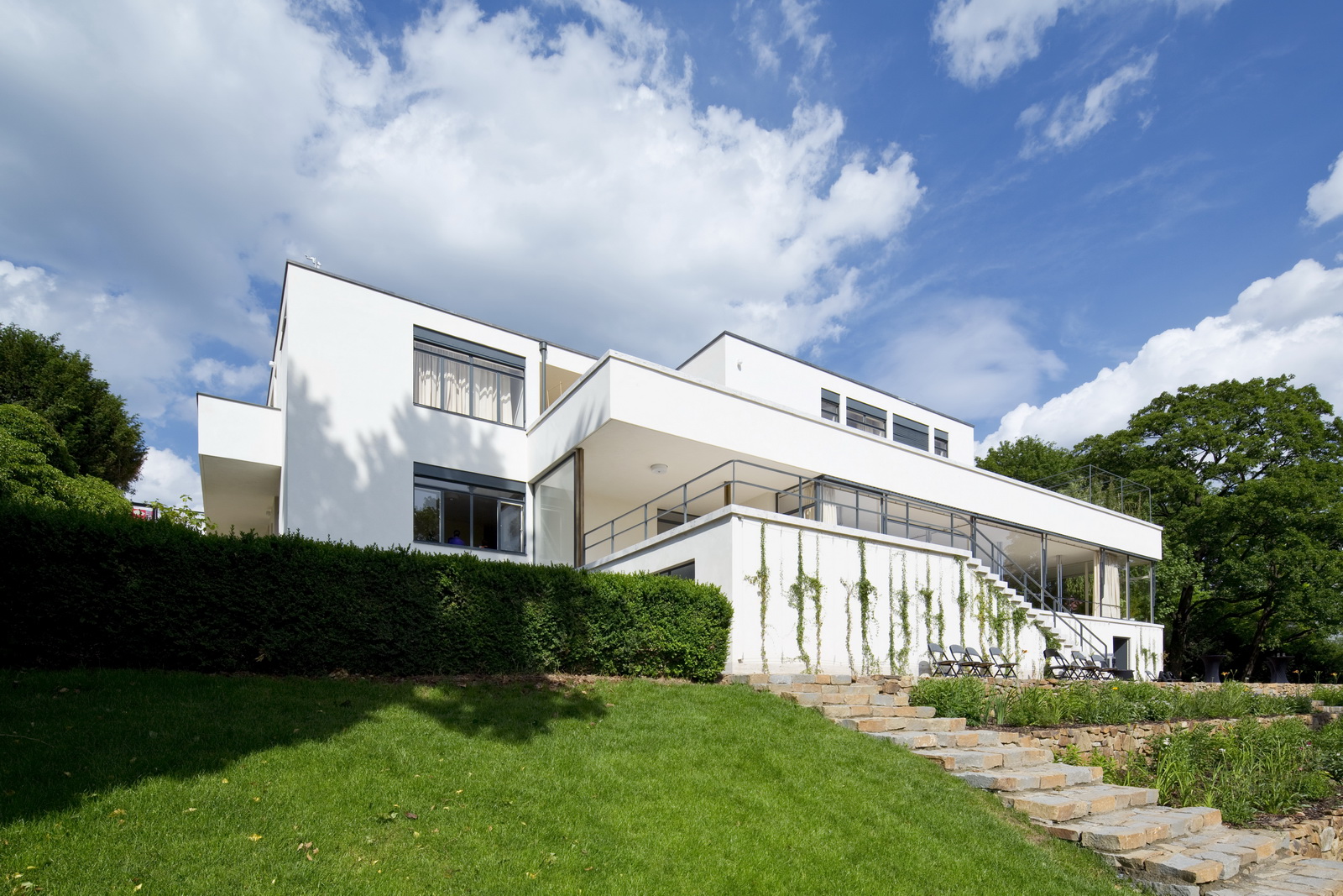 Moderná biela vila obklopená zelenou záhradou pod modrou oblohou s oblakmi.