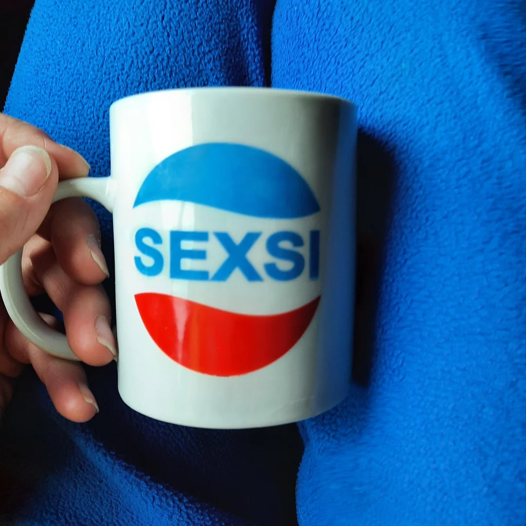 Ruka drží bielu hrnček s nápisom SEXSI, modročervená potlač, modrý textil v pozadí.