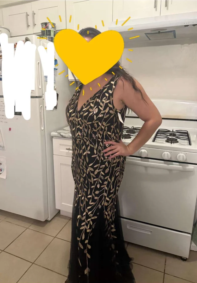 Osoba v kuchyni v čiernych šatách s listovým vzorom, rúra a chladnička v pozadí.