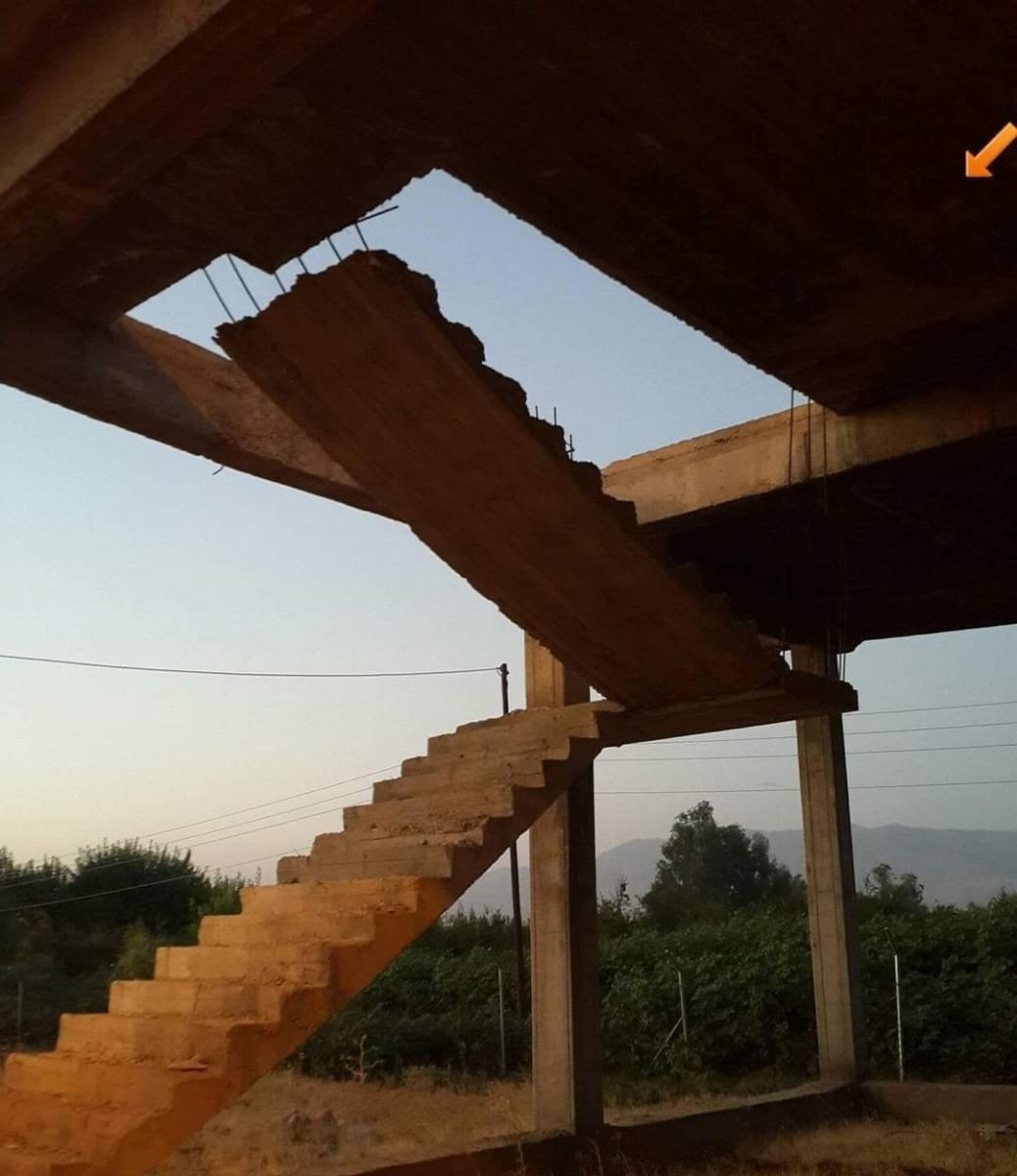 Nedokončené betónové schodisko v exteriéri budovy bez strechy, s výhľadom na prírodu a kopce v pozadí.