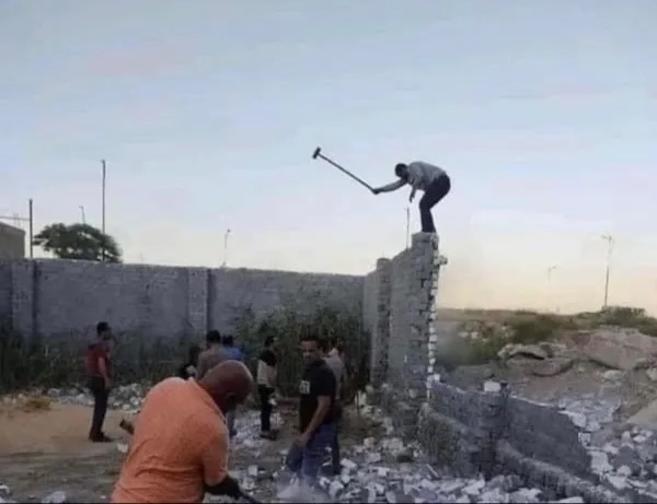 Skupina mužov demoluje tehlový múr, jeden z nich pracuje zhora pomocou kladiva.