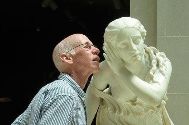 Man listens closely to a statue of a woman in a playful interaction.