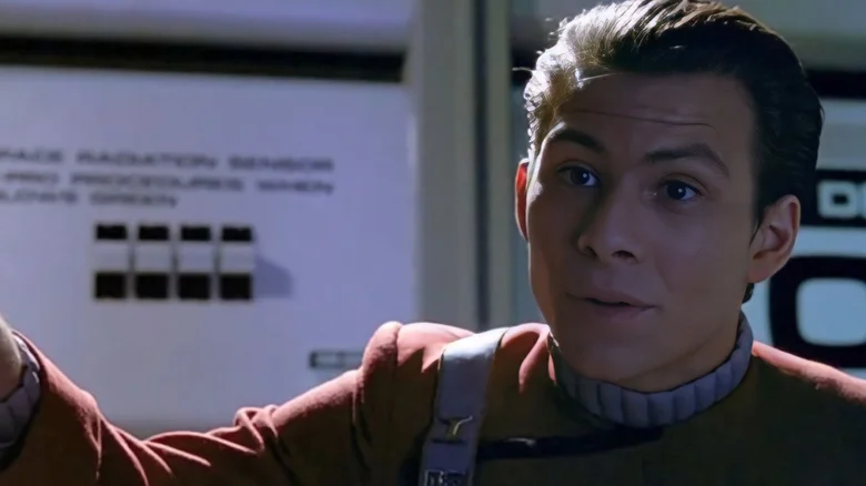 Muž v sci-fi uniforme hovorí na vesmírnej lodi, pozadie s panelmi.