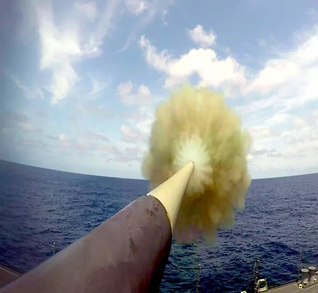 Delo vypúšťa projektil nad oceánom, zachytené v momente výstrelu, modrá obloha a oblaky na pozadí.