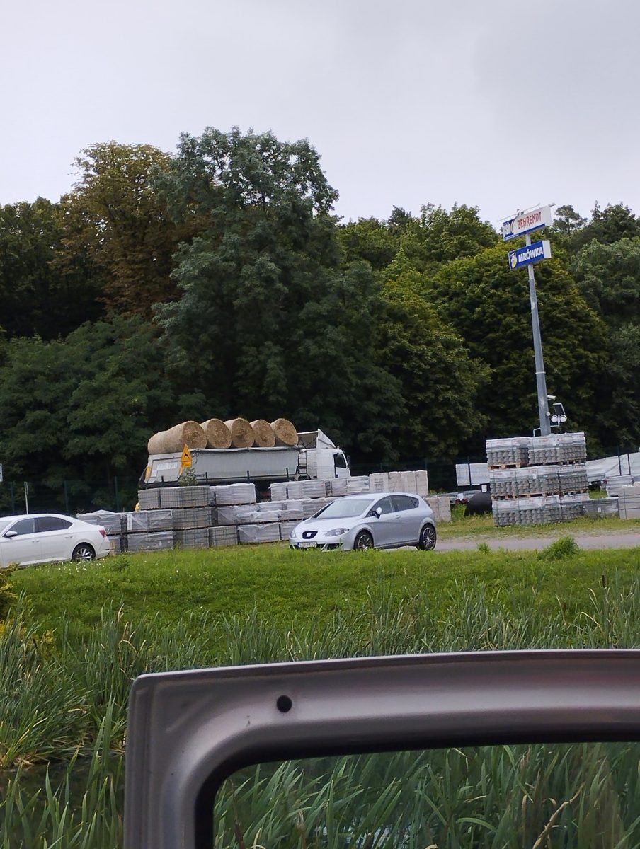 Kamión prevážajúci balíky sena, autá na parkovisku pred lesom, sklad stavebného materiálu na pozadí.