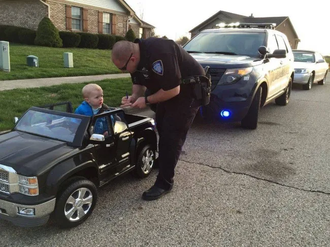 Dieťa na hračkárskom aute zastavené policajtom na sídlisku, vedľa nich stojí policajné auto.