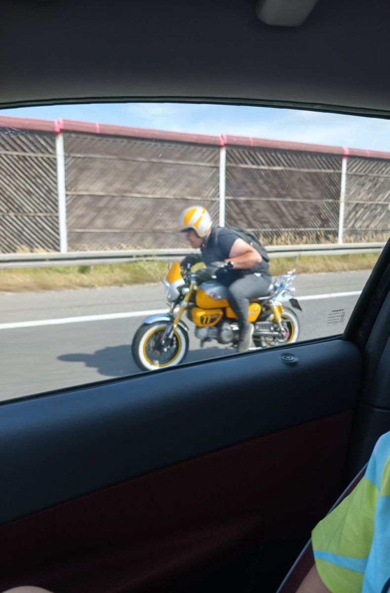 Motorkár v žltom prilbe jazdí na malom motocykli po diaľnici za slnečného dňa.