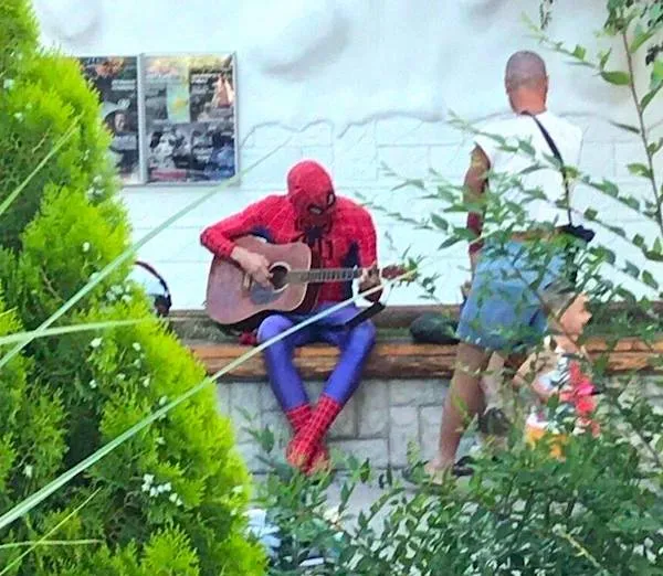 Muž v kostýme hrá na gitare na lavičke v parku, popri ňom prechádzajú okoloidúci.