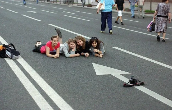 Skupina ľudí ležiacich na ceste s fotoaparátom, pripravení na kreatívne fotenie.