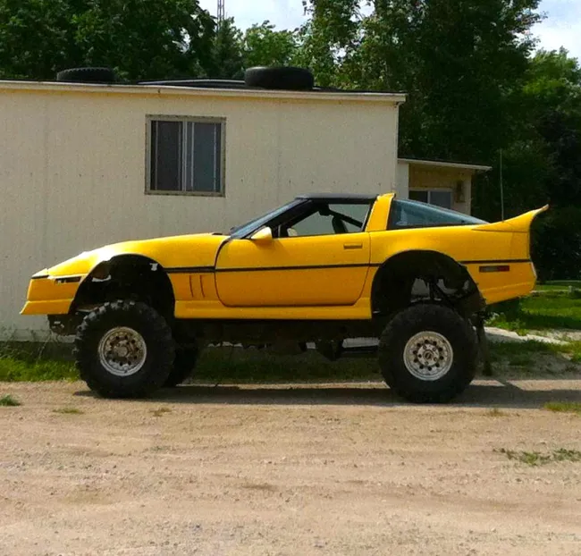 Žlté auto so zvýšeným podvozkom, zaparkované pred bielou budovou, obklopené zelenými stromami.