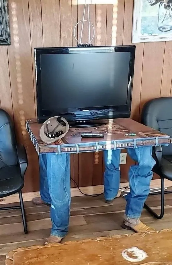 TV na stene so stolíkom v tvare nôh s džínsami a topánkami, klobúk položený na stole.