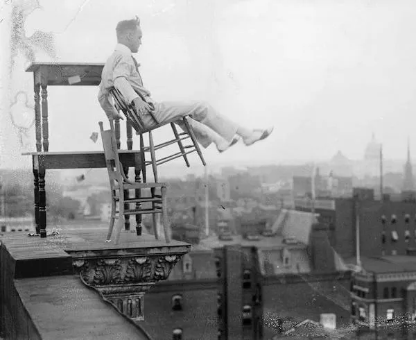 Muž sediaci na stoličke na kraji strechy nad mestom, odrážajúci odvážnu akrobaciu.