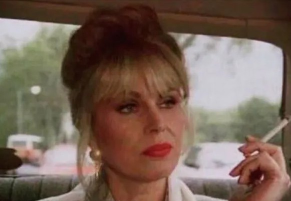 Žena s rúžom a cigaretou sedí v aute, pohľad smeruje von z okna. Sústredený výraz tváre.