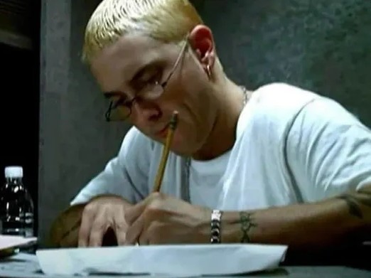 Osoba s blond vlasmi a okuliarmi píše na papier, obklopená tmavým prostredím a fľašou vody na stole.