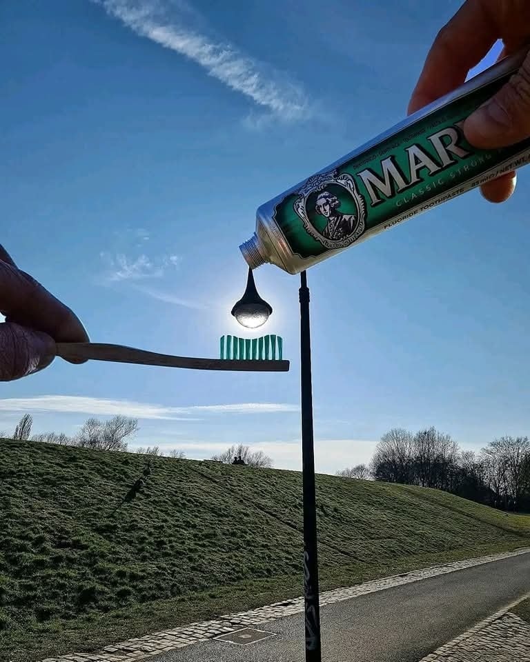 Osoba používa perspektívu na ilúziu, že vytláča slnko z tuby na zubnú kefku, s jasnou oblohou v pozadí.