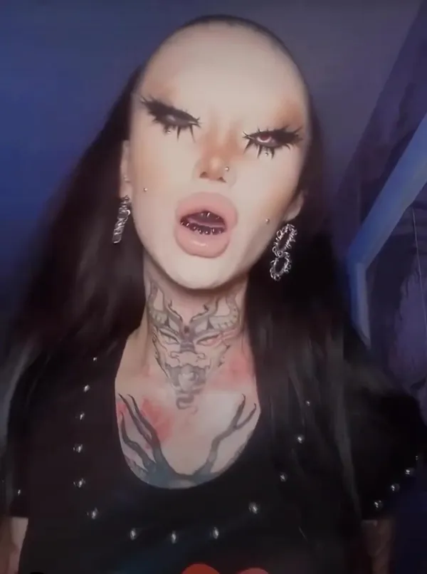 Osoba s výrazným líčením, piercingami a tetovaním na krku v tmavom prostredí.