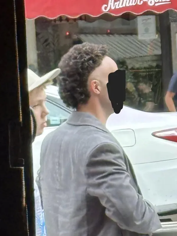 Osoba so zaujímavým účesom stojí na ulici vedľa auta, v pozadí červená markíza.