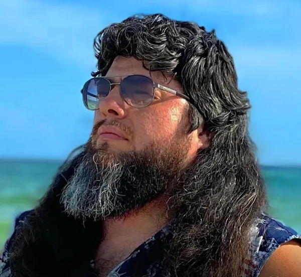 Osoba s dlhými vlasmi a okuliarmi na pláži, pohľad na jasnú oblohu a more v pozadí.