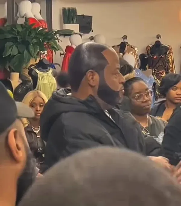 Skupina ľudí v obchode s oblečením, muž v popredí má výrazný účes a upravenú bradu.