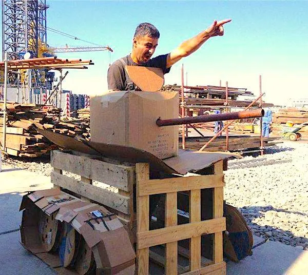 Muž na stavbe v improvizovanom tanku z kartónu a dreva gestikuluje rukou do diaľky.