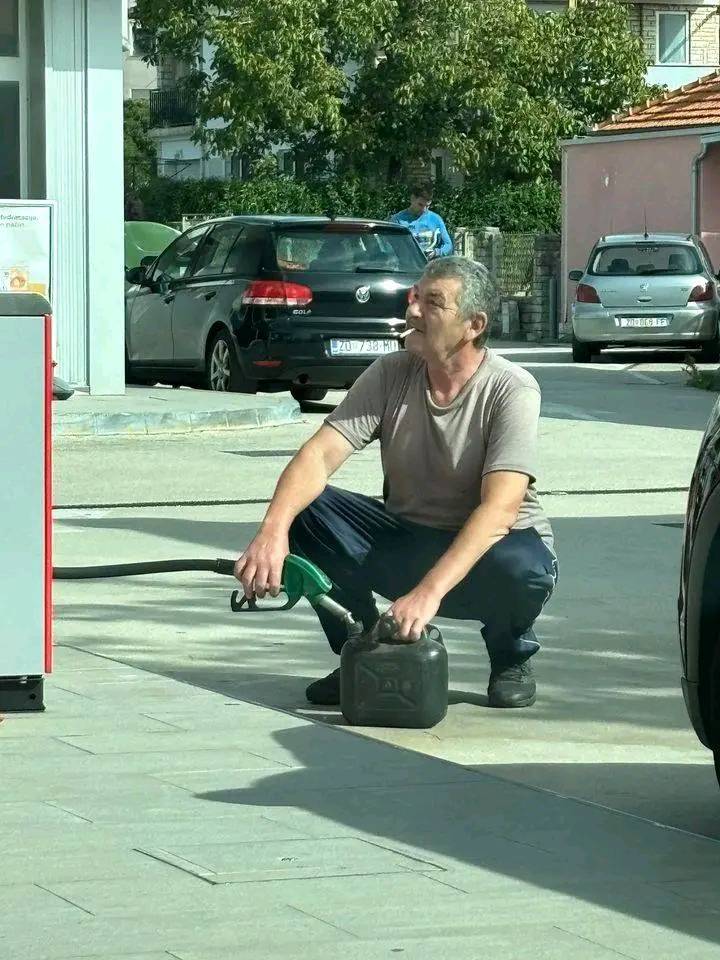 Muž na čerpacej stanici plní kanystr palivom, sedí v podrepe vedľa auta na parkovisku.