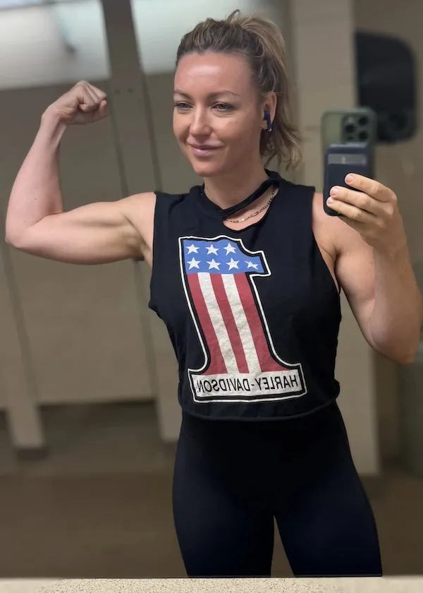 Osoba v posilňovni fotografuje svoj biceps, oblečená v čiernom tričku s americkou vlajkou a nápisom Harley-Davidson.