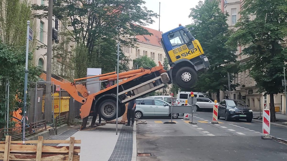 Nákladné auto sa nakláňa na mestskom stavenisku, zdvíha sa prednou časťou, zatiaľ čo pracovníci sledujú situáciu.