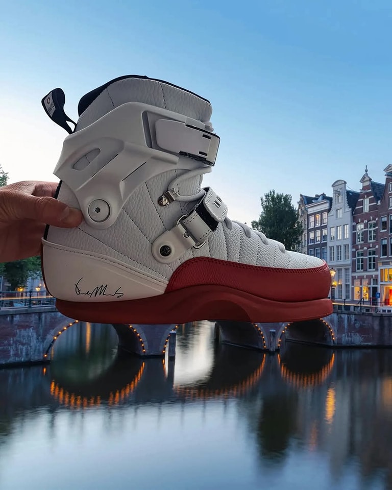 Biela športová topánka s červenou podrážkou, držiaca pred vodným kanálom a historickými budovami v pozadí.