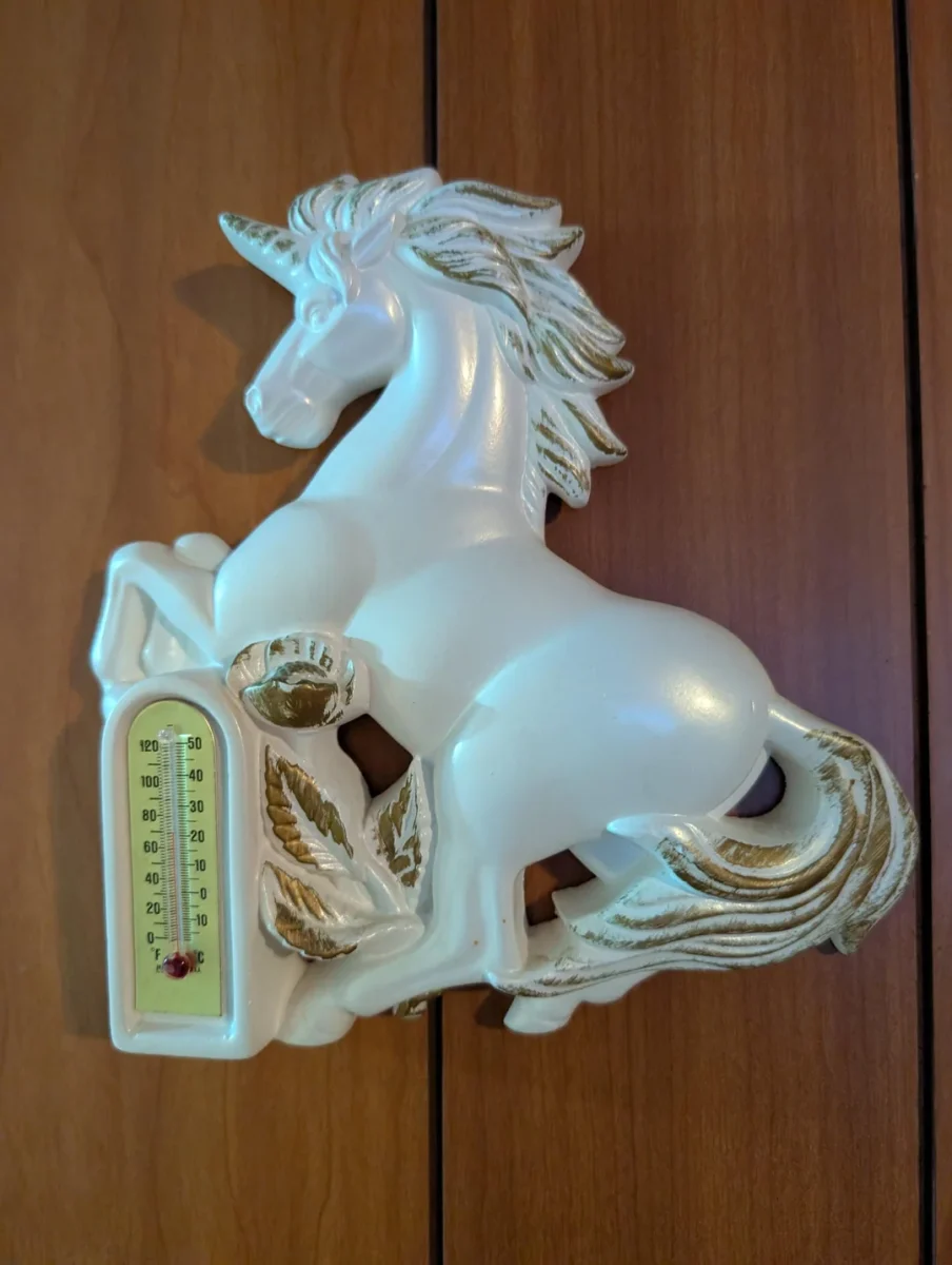 Biely keramický jednorožec s teplomerom a zlatými detailmi na drevenej stene.