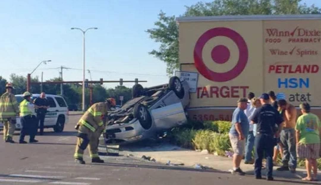 Auto prevrátené na streche pri billboarde s logom, záchranári a ľudia na mieste nehody.