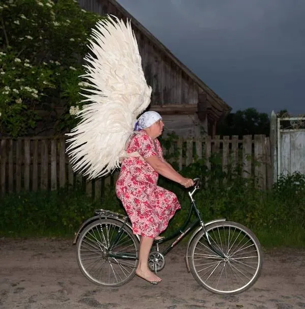 Žena v šatách s obrovskými krídlami jazdí na bicykli pred drevenou chalupou za súmraku.
