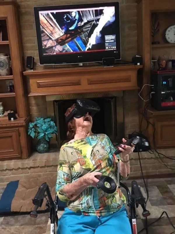 Osoba sediaca na vozíku užíva virtuálnu realitu s náhlavnou súpravou a ovládačmi, zatiaľ čo na televízore beží hra.