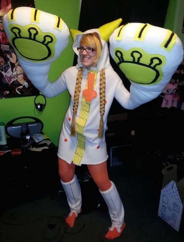 Osoba v zábavnom kostýme zvieraťa s veľkými rukavicami, dlhými vrkočmi, stojaca v izbe s farebnou stenou.