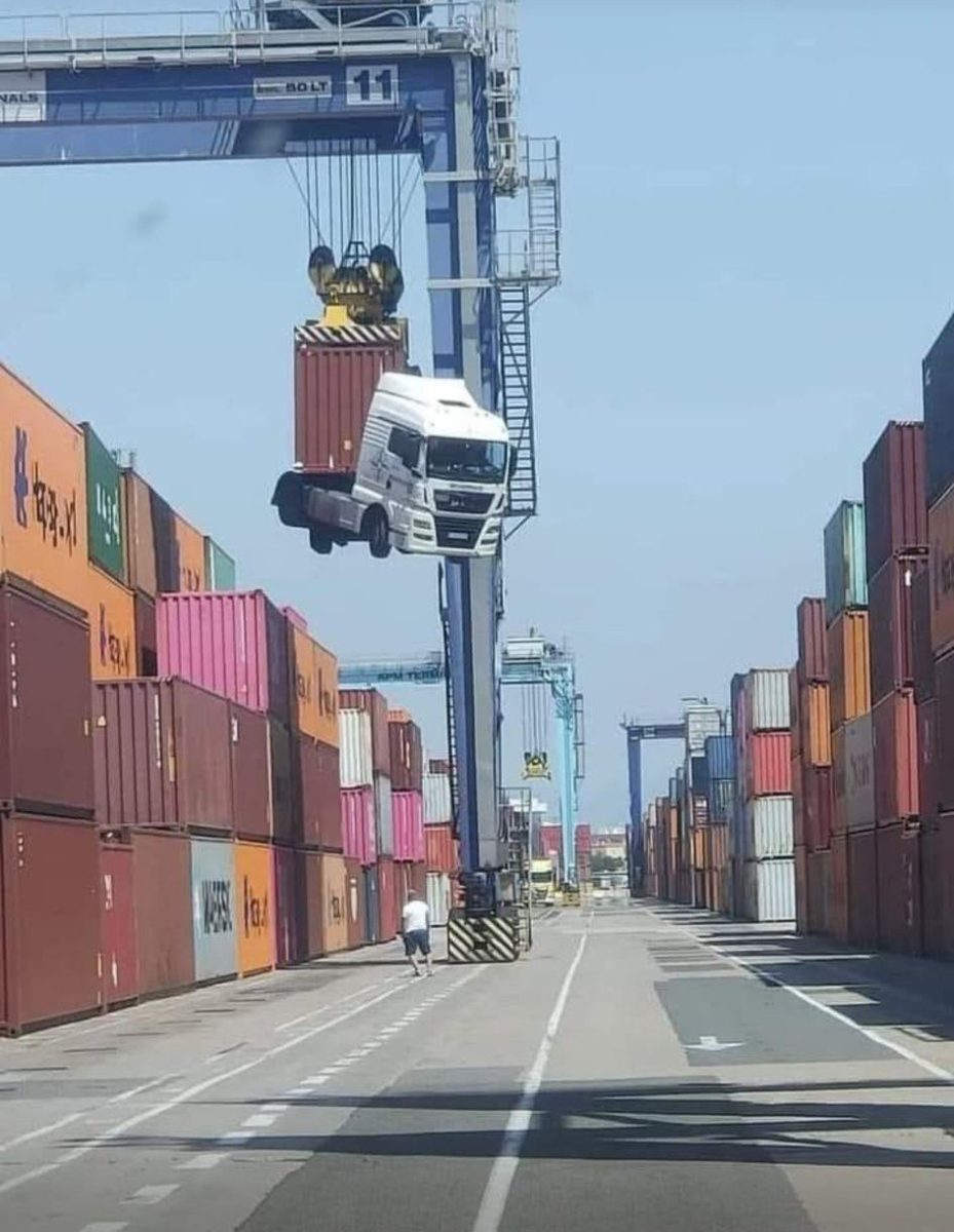 Nákladný automobil je zdvihnutý žeriavom nad prístavným priestorom plným kontajnerov.