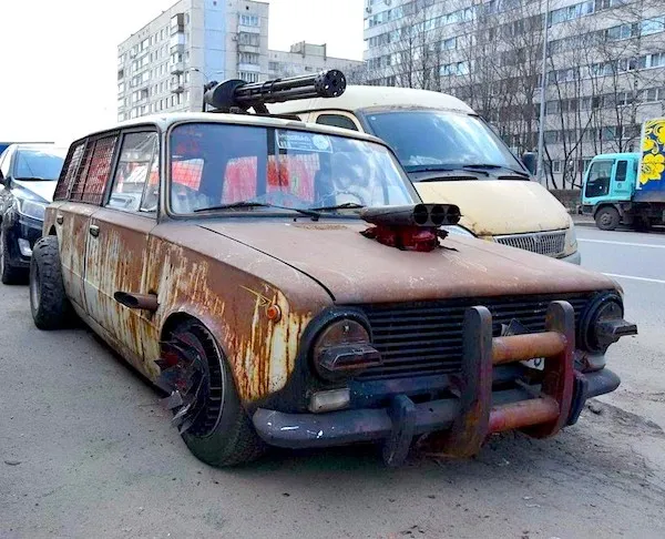 Ošarpané auto s drsným vojenským dizajnom, parkujúce na ulici vedľa iných vozidiel.