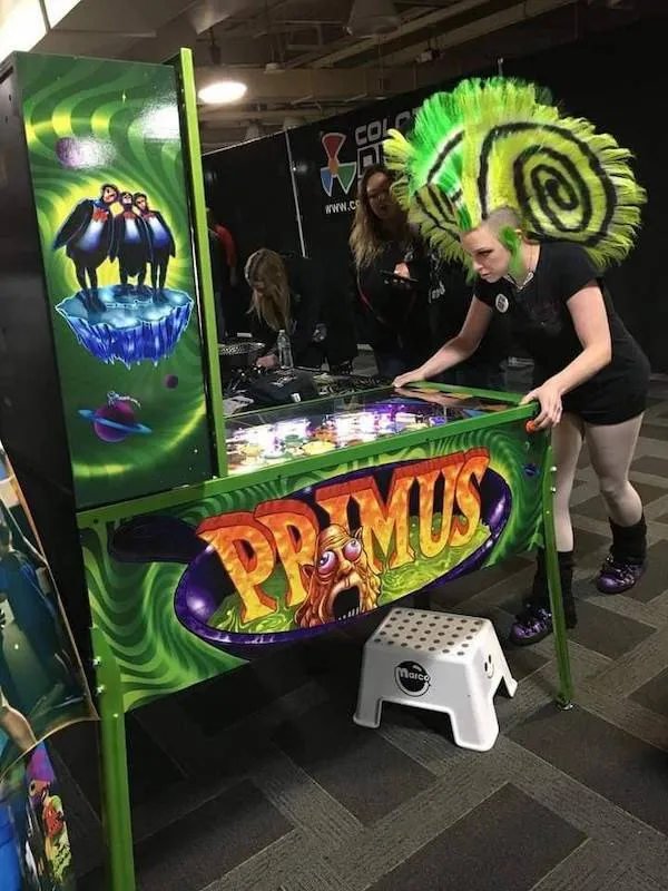 Osoba so zeleným čírom hrá na farebnom pinballe s nápisom Primus v interiéri herne.