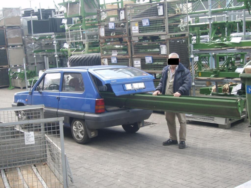 Muž nakladá dlhý kovový nosník do malého modrého auta na parkovisku plnom priemyselného vybavenia.