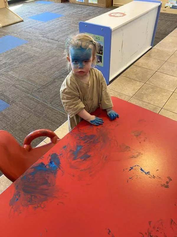 Dieťa s modrou tvárou a rukami pri červenom stole v farebnej miestnosti.