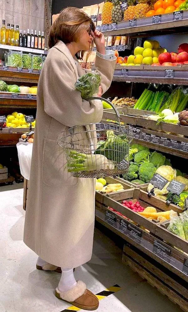 Žena v kabáte nakupuje zeleninu v obchode s potravinami; drží kôš plný čerstvých produktov.