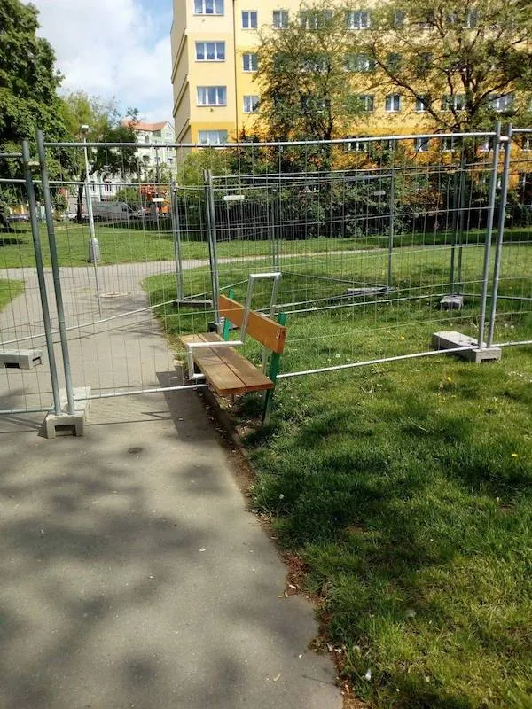 Lavička za plotom na trávniku vedľa chodníka, v pozadí žltá budova a stromy.