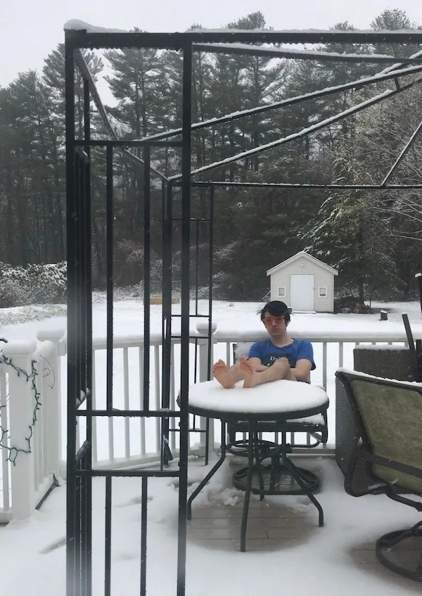 Osoba sedí na zasneženom dvore, s nohami na stole, obklopená zimnou krajinou a zimnou výzdobou.