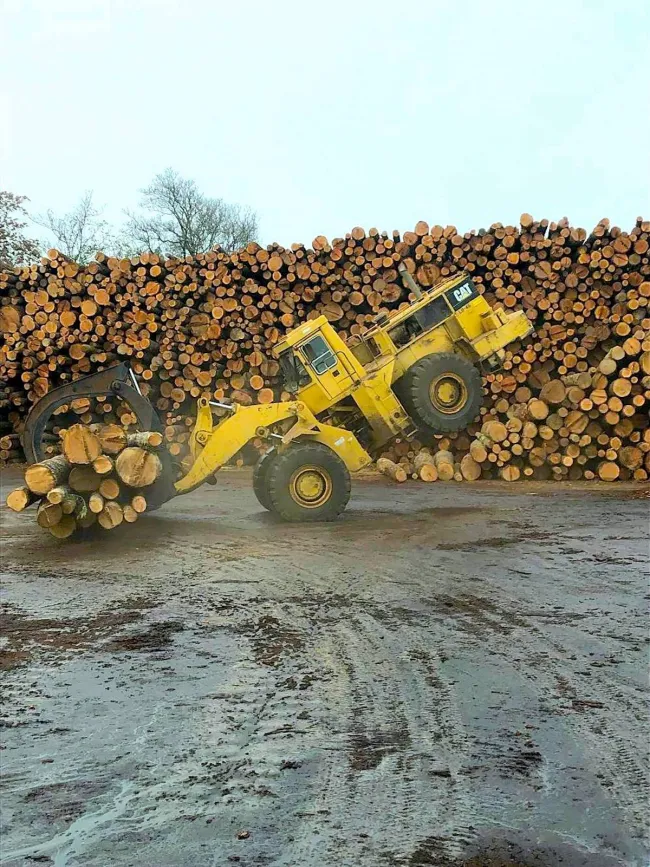 Žltý nakladač s drapákom zdvíha veľké kmene stromov pred štosom narezaného dreva vo vonkajšom priestore píly.