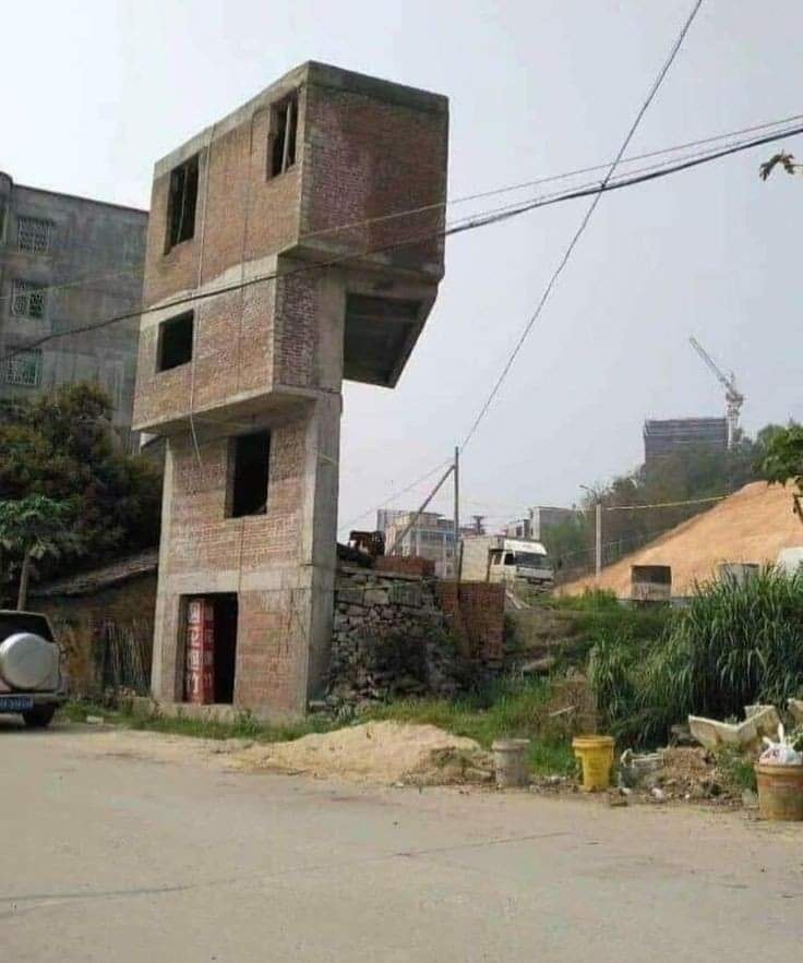 Neobvyklá nedokončená trojposchodová budova z tehál na vidieku, vyzerajúca nestabilne a asymetricky postavená.