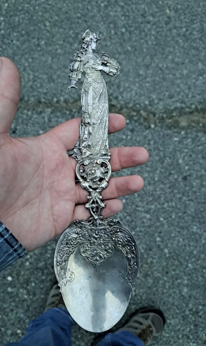 Dekoratívna strieborná lyžica s detailným reliéfom postavy v ruke, položená na betóne.