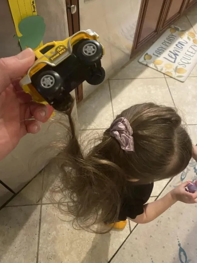 Detské autíčko zapletené v dievčenskom cope, zábavná nehoda na dlažbe kuchyne.