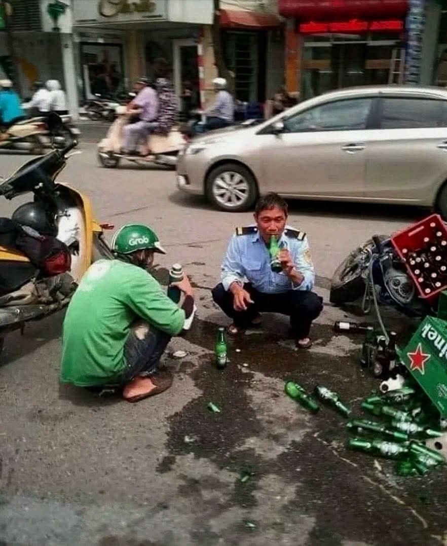 Dvaja muži na ulici s prevrátenými fľašami piva, okolité vozidlá a chodec na pozadí.
