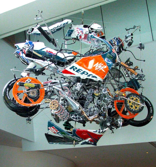 Závesná inštalácia rozloženej motorky s oranžovými, čiernymi a bielymi prvkami vo vzduchu v modernom priestore.