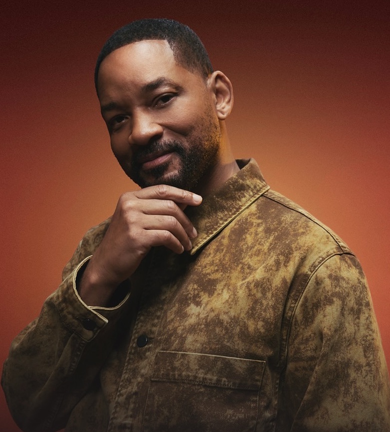 Osoba v hnedých šatách s rukou na brade, póza pred oranžovým pozadím.